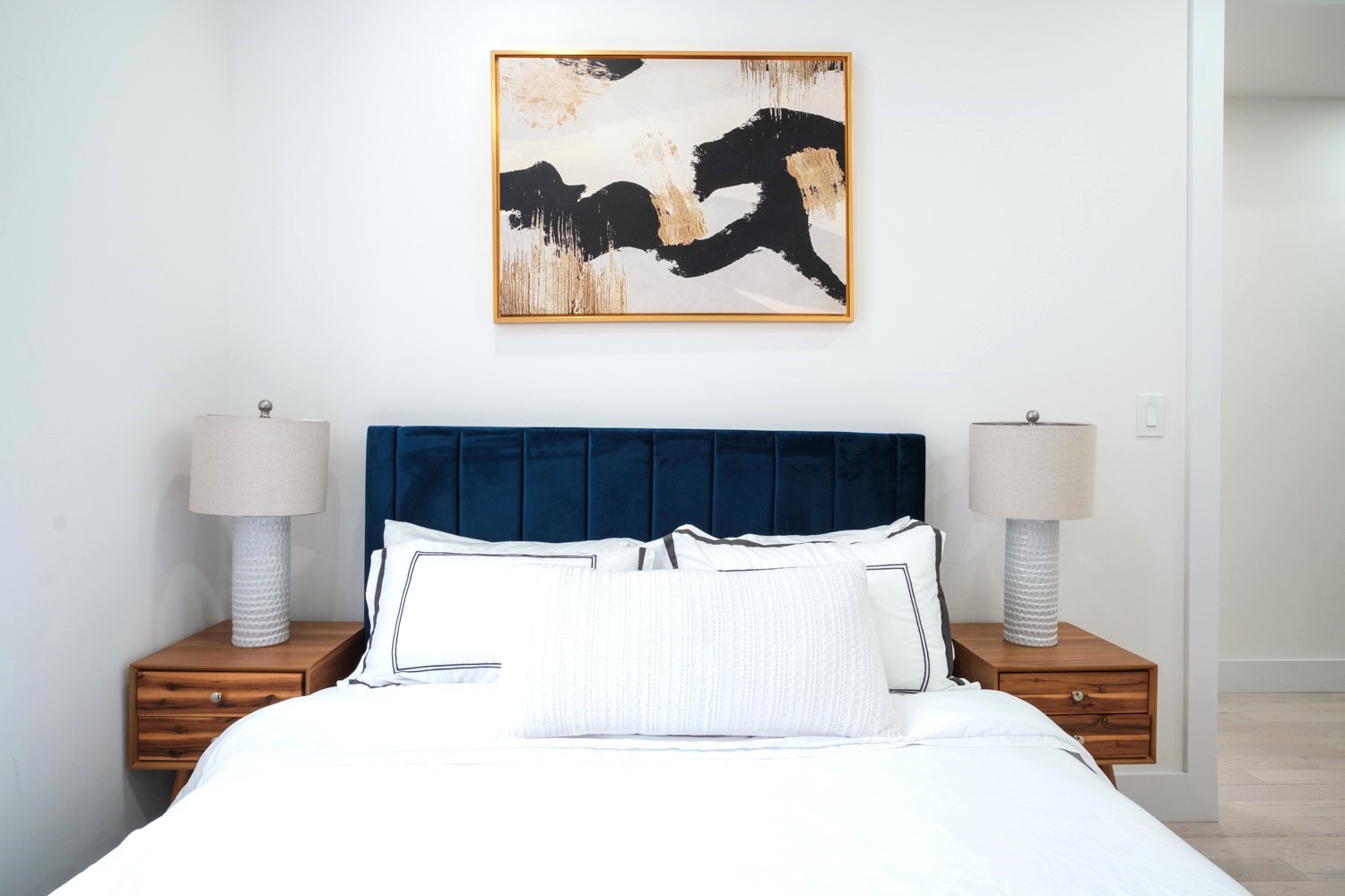 Bedroom with navy velvet headboard, walnut nightstands and gold framed abstract art